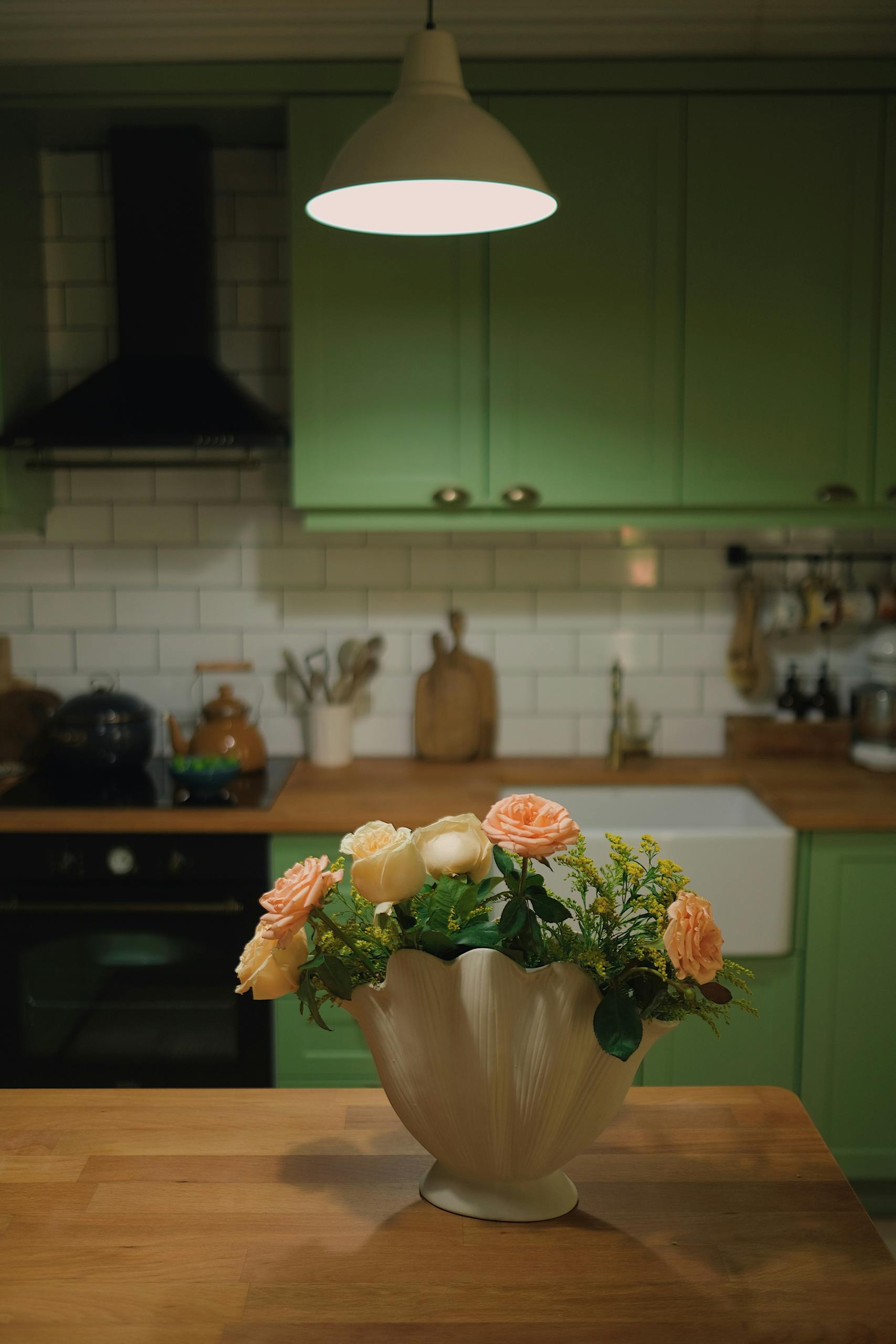 A cozy kitchen interior with a floral arrangement under warm lighting, featuring pastel green cabinets.