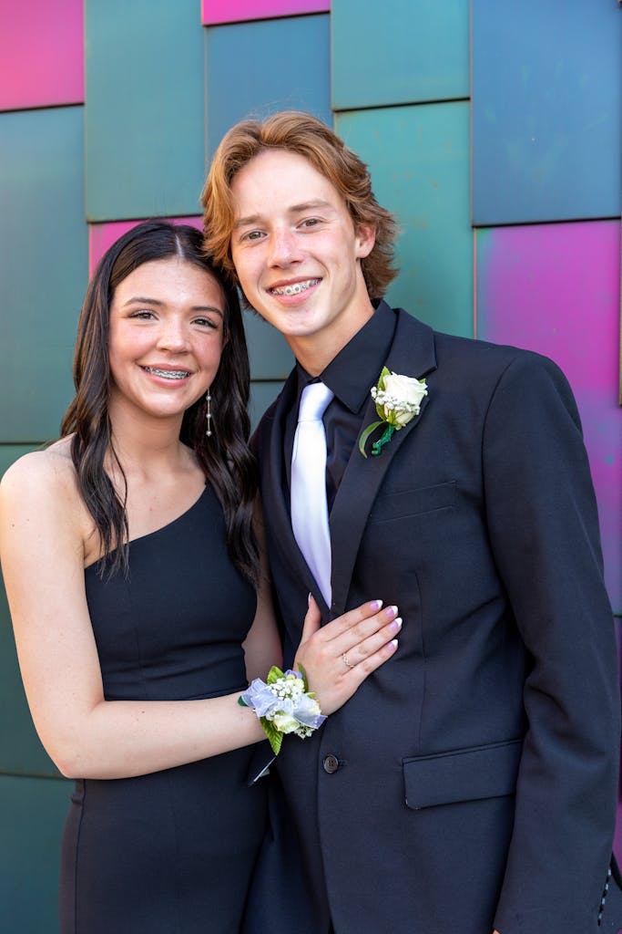 Stylish young couple in formal attire posing outdoors against a colorful backdrop.